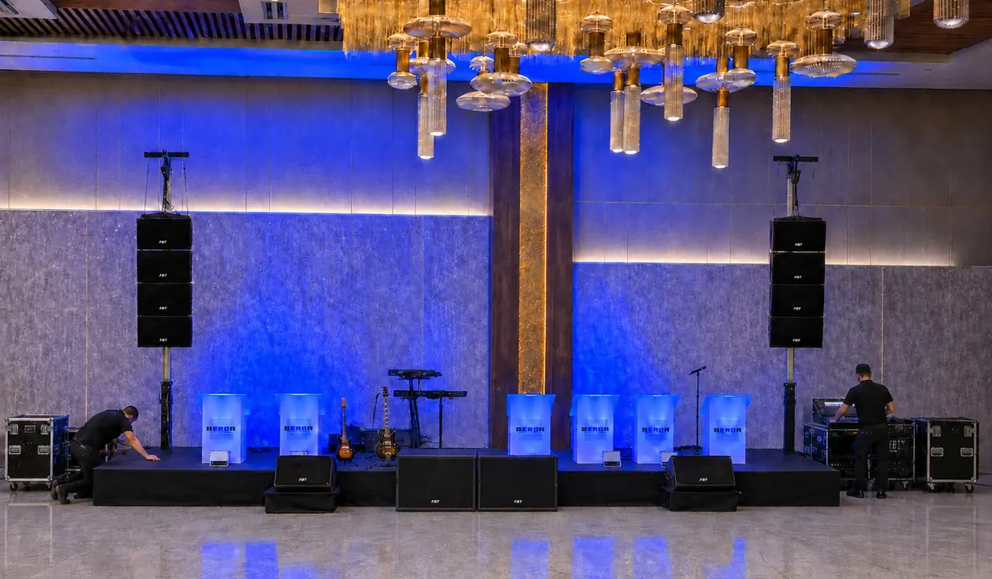 Ballroom stage set with flown line-array speakers, floor subwoofers, stage monitors, band instruments, and blue uplighting while two technicians finish the setup