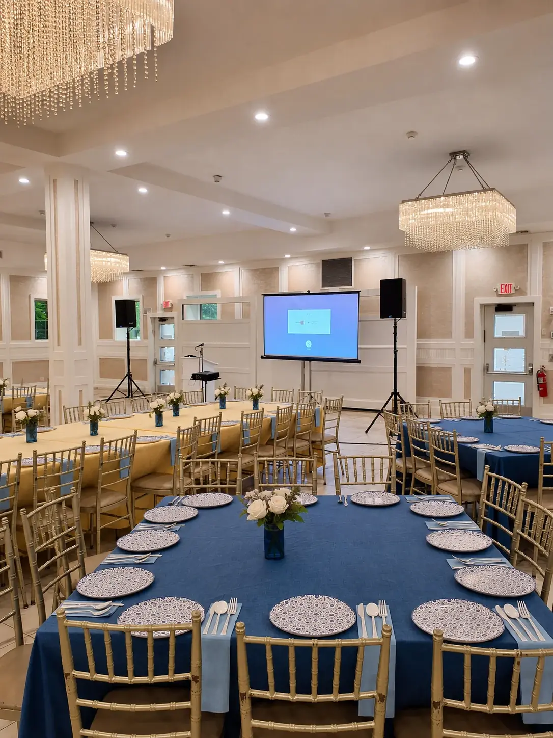 Banquet room with long dressed tables and place settings facing a projection screen flanked by two speakers on stands, with a microphone at a small lectern
