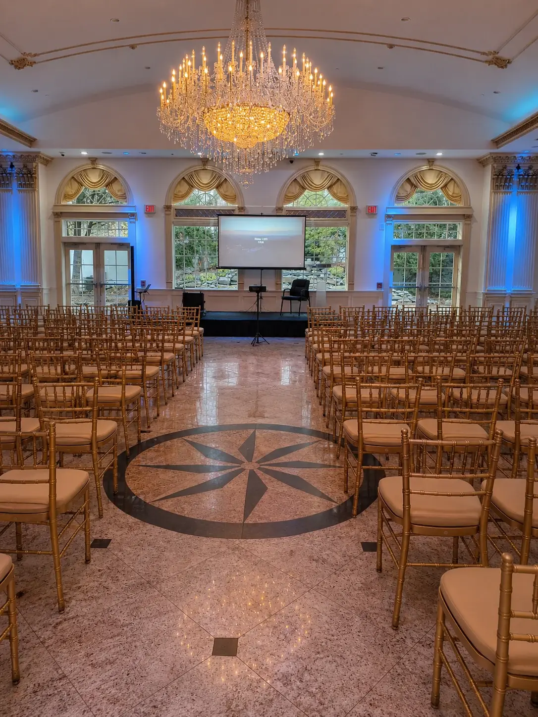 Formal hall seated in rows of gold chairs facing a projection screen on a tripod with a projector on a stand and blue wall uplighting