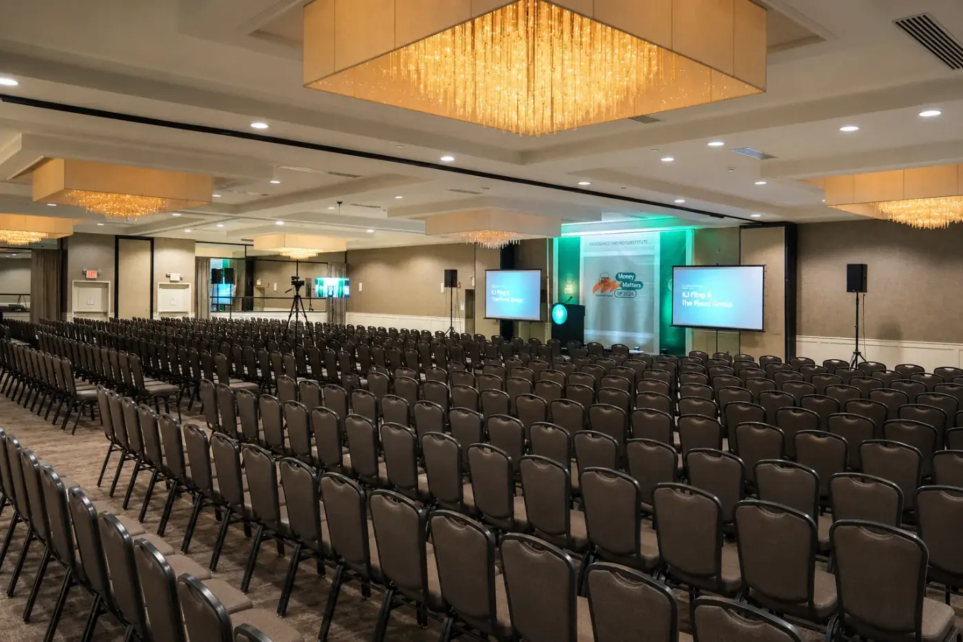 Large conference room seated for hundreds facing two projection screens, a lectern, and speakers on stands, with a camera on a tripod at the back