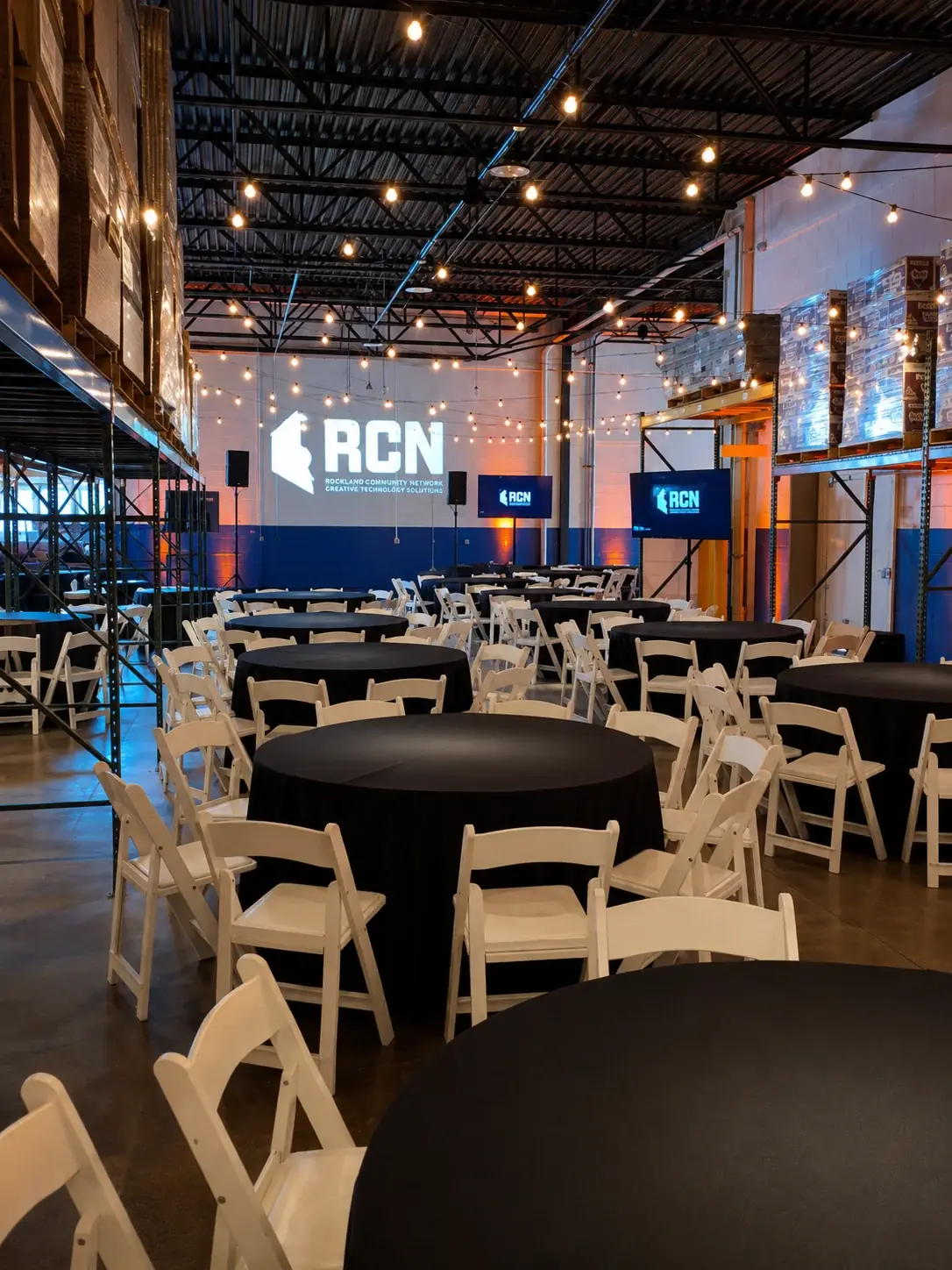 Warehouse venue set with round tables and white folding chairs, string lighting overhead, a large projected logo on the back wall, and two flat-panel displays on stands